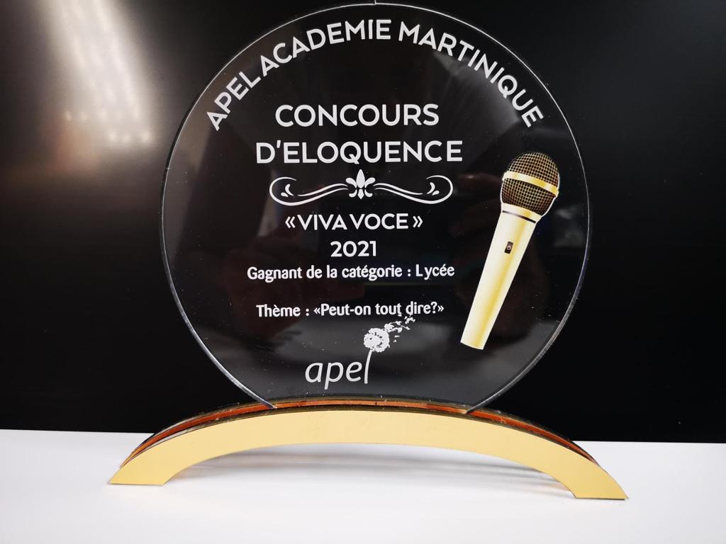 Trophée en verre transparent avec un micro doré sur fond noir. Texte : « APELACADÉMIE MARTINIQUE, Concours d'éloquence, Viva Voce 2021, Lycée, Peut-on tout dire ? »