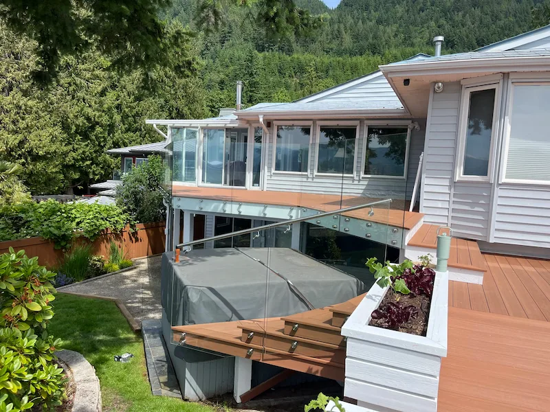 Multi-level backyard deck renovation ideas featuring frameless glass railings, wood composite decking, built-in planter box, and covered lower patio surrounded by lush greenery.