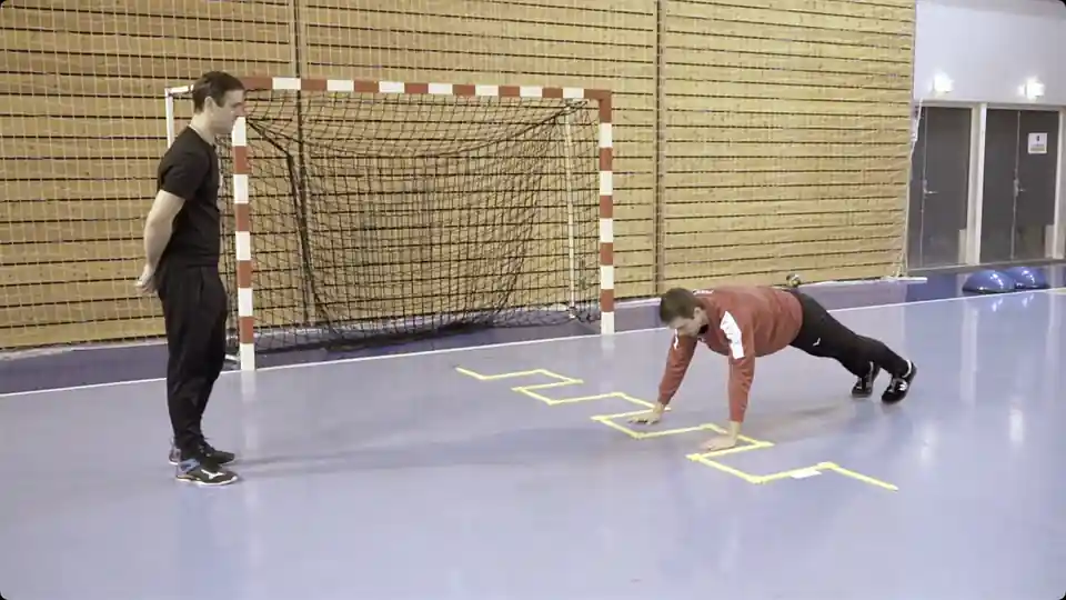 A coach watches a handball goalkeeper in a red top perform an agility-ladder warm-up with hands on the court in front of the goal.