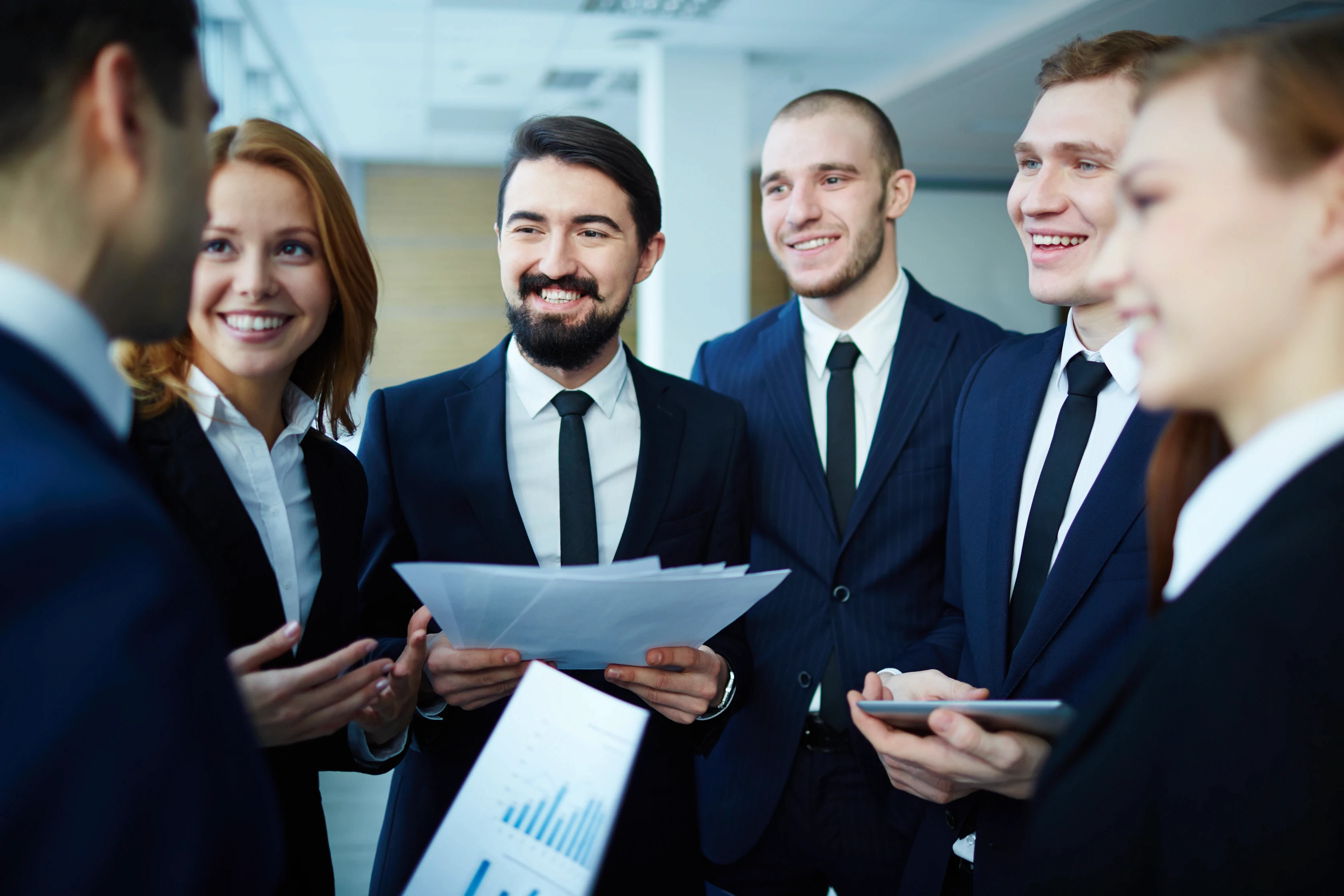 A group of five business professionals in suits are smiling and engaged in a discussion. One holds documents, while another holds a tablet, suggesting teamwork.