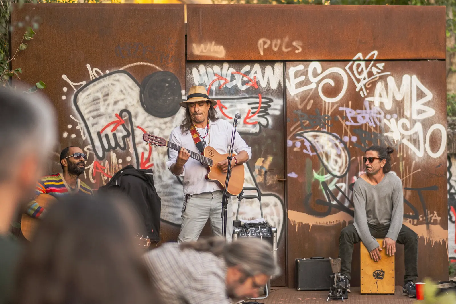 A street performer plays music on stage while an audience watches against a colorful mural backdrop.