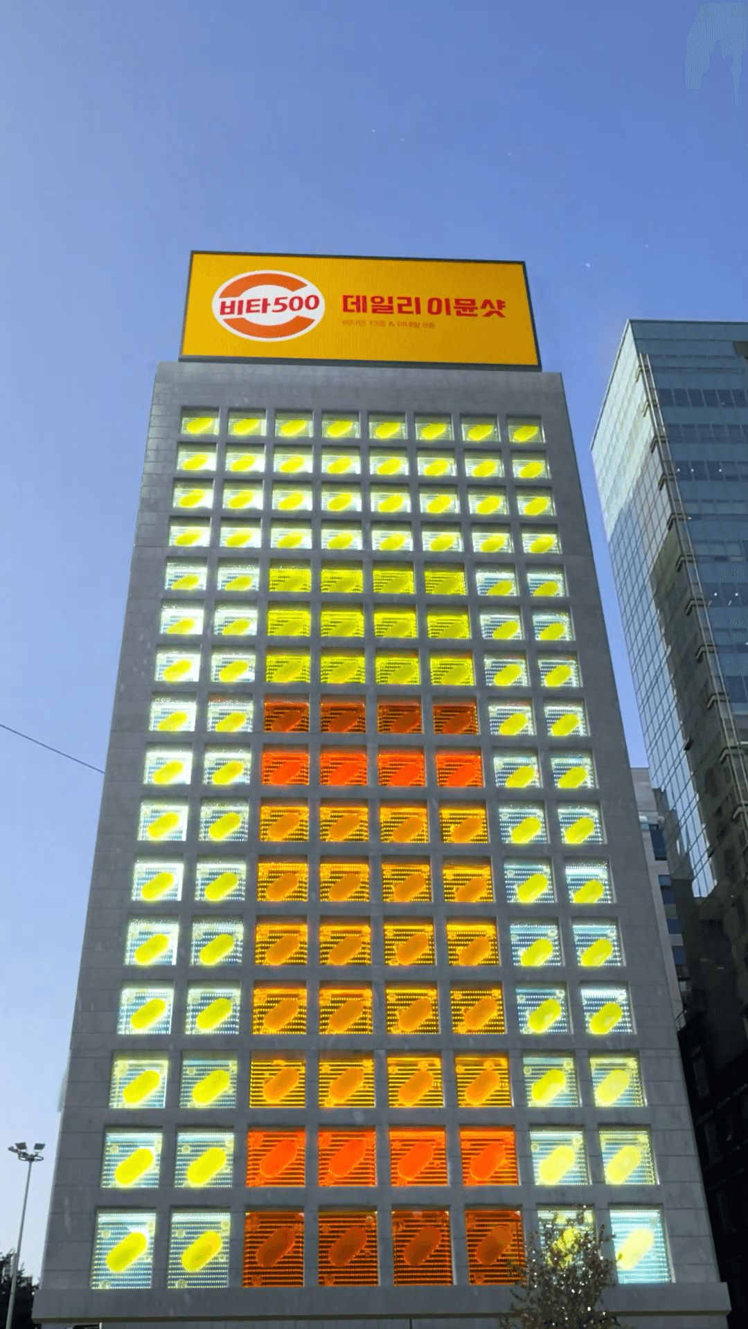 노란색과 주황색 비타민 캡슐 블록이 테트리스처럼 건물 외벽을 가득 채운 FOOH 장면