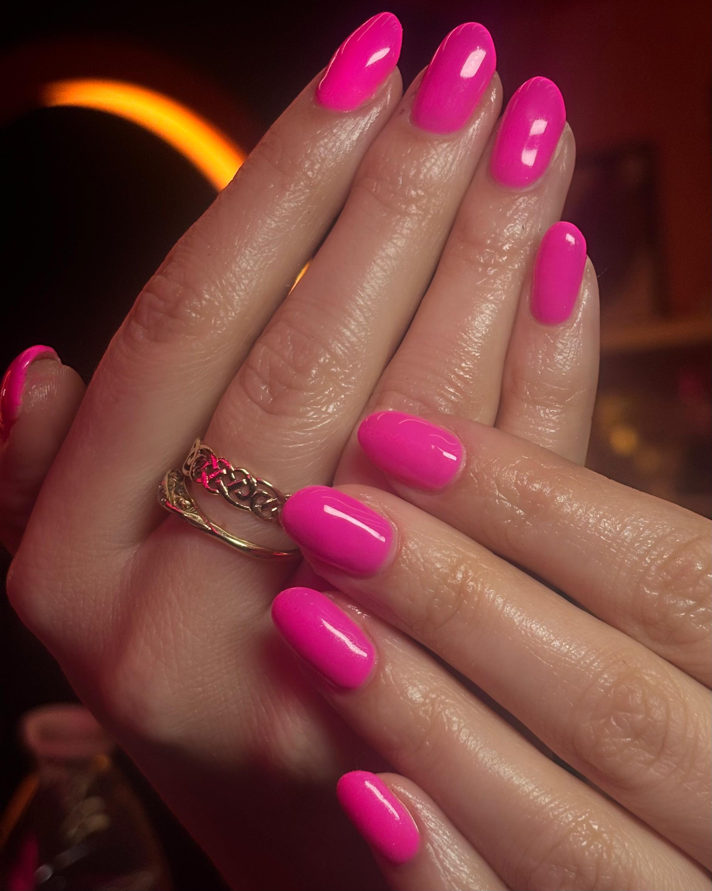 Short, oval-shaped nails in a solid, high-gloss "Barbie" pink. The finish is extremely reflective, shown under warm ring lighting with a gold Celtic-style ring on the finger.