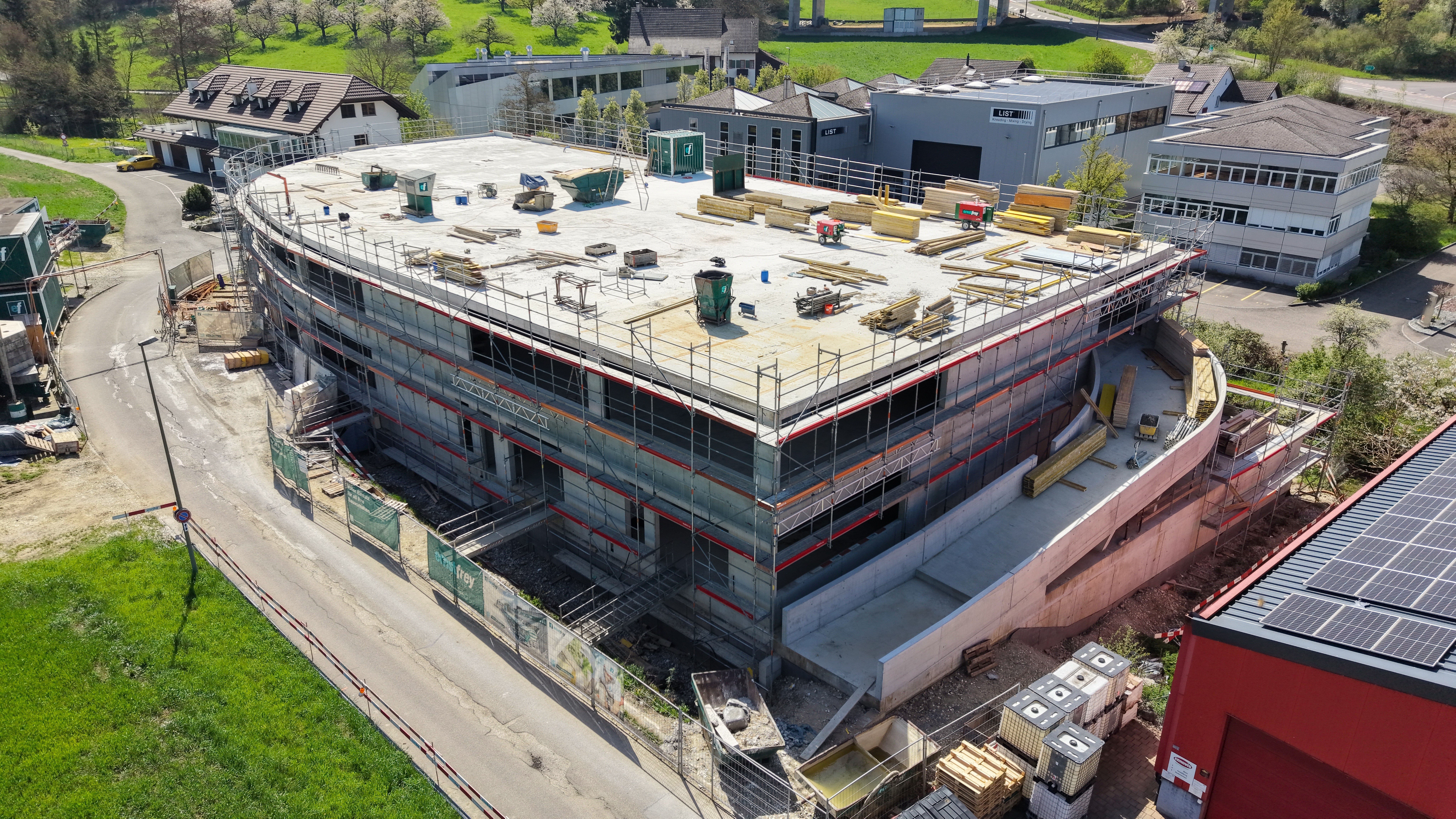 Baustelle eines modernen Gewerbegebäudes mit Rohbau, Gerüst und umliegender Infrastruktur aus der Luftperspektive
