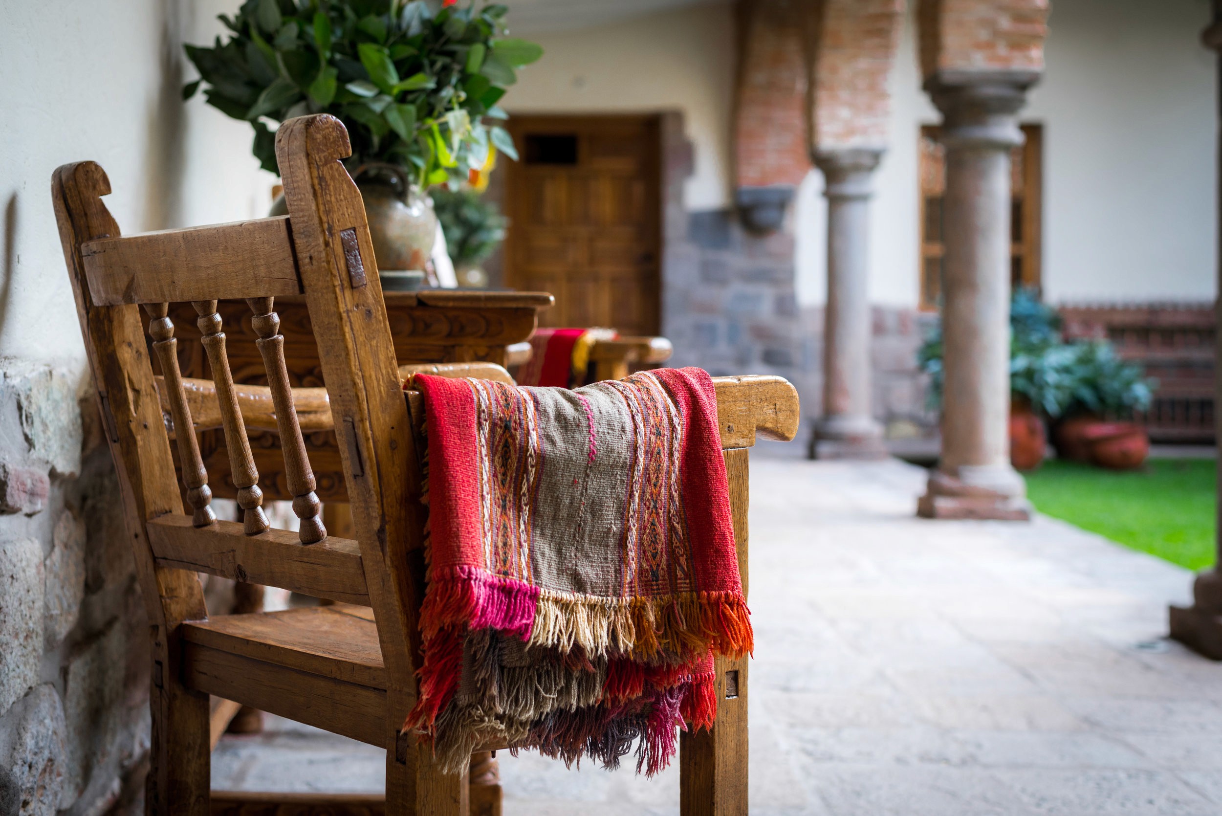 A wooden chair with a colorful striped woven blanket draped over it sits on a stone patio near an arched stone doorway.