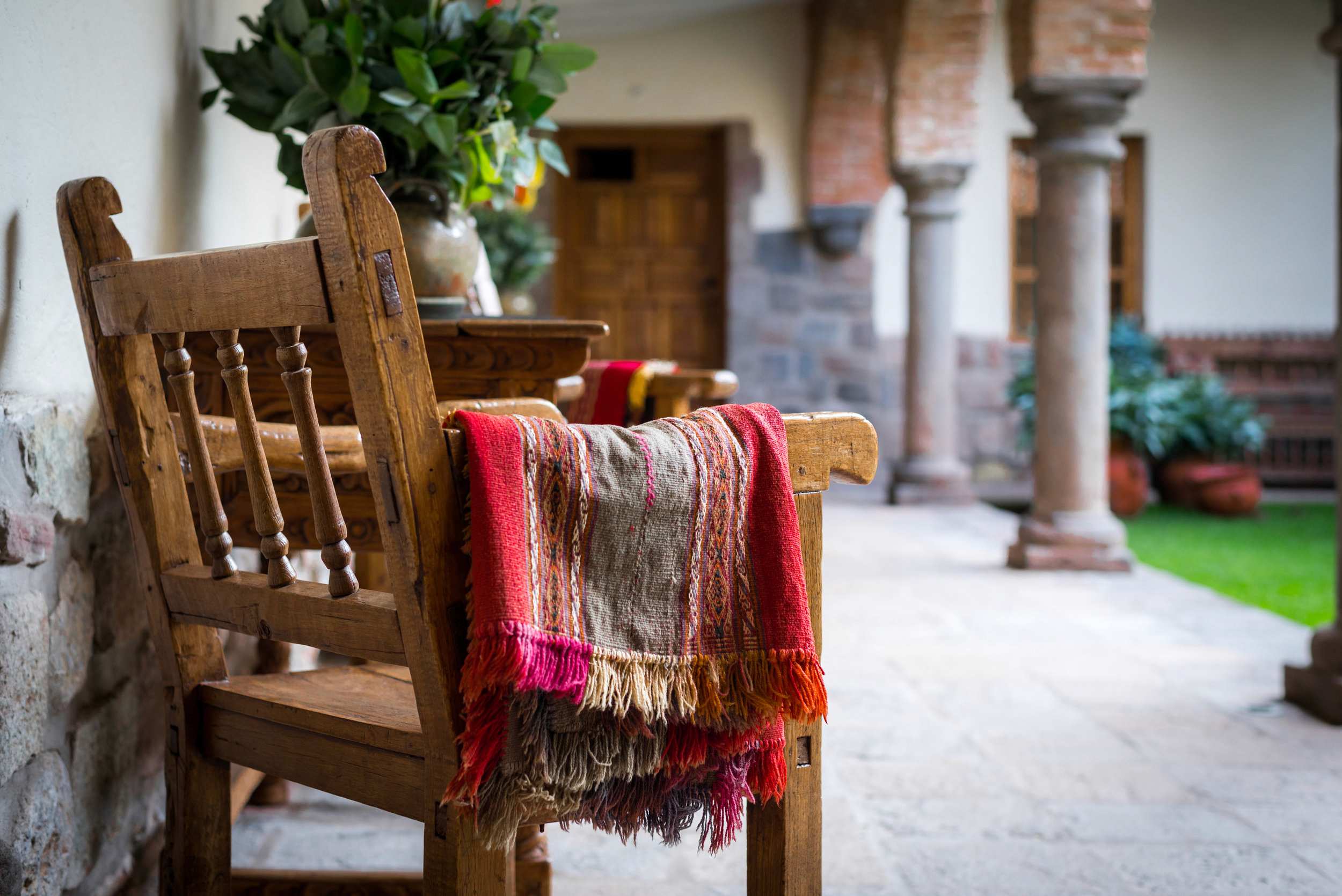 A wooden chair with a colorful striped woven blanket draped over it sits on a stone patio near an arched stone doorway.