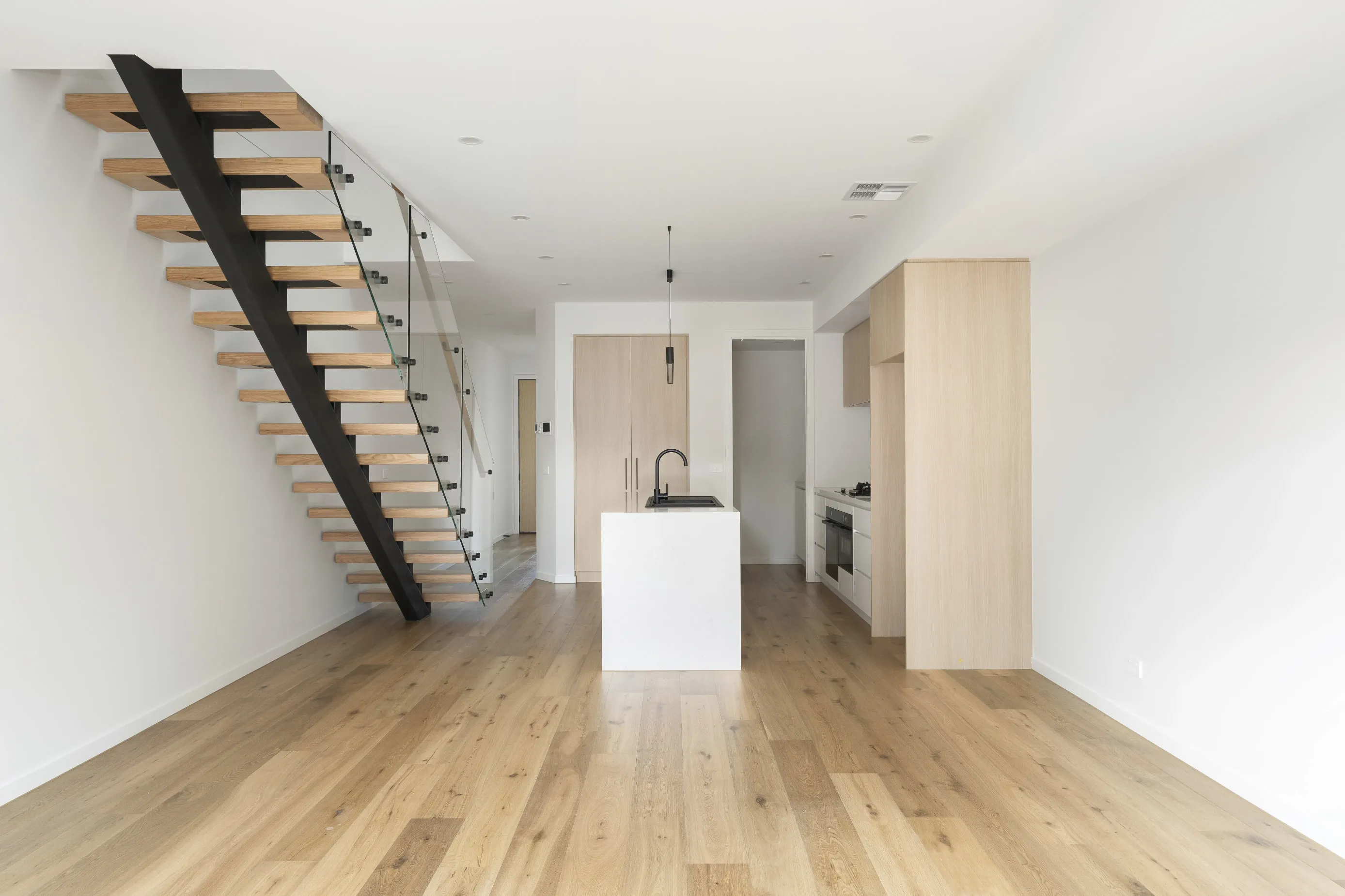 Open living and kitchen with white walls, oak floors, a floating timber staircase with black steel stringer and glass balustrade and a white-and-oak island with black tap and pendant