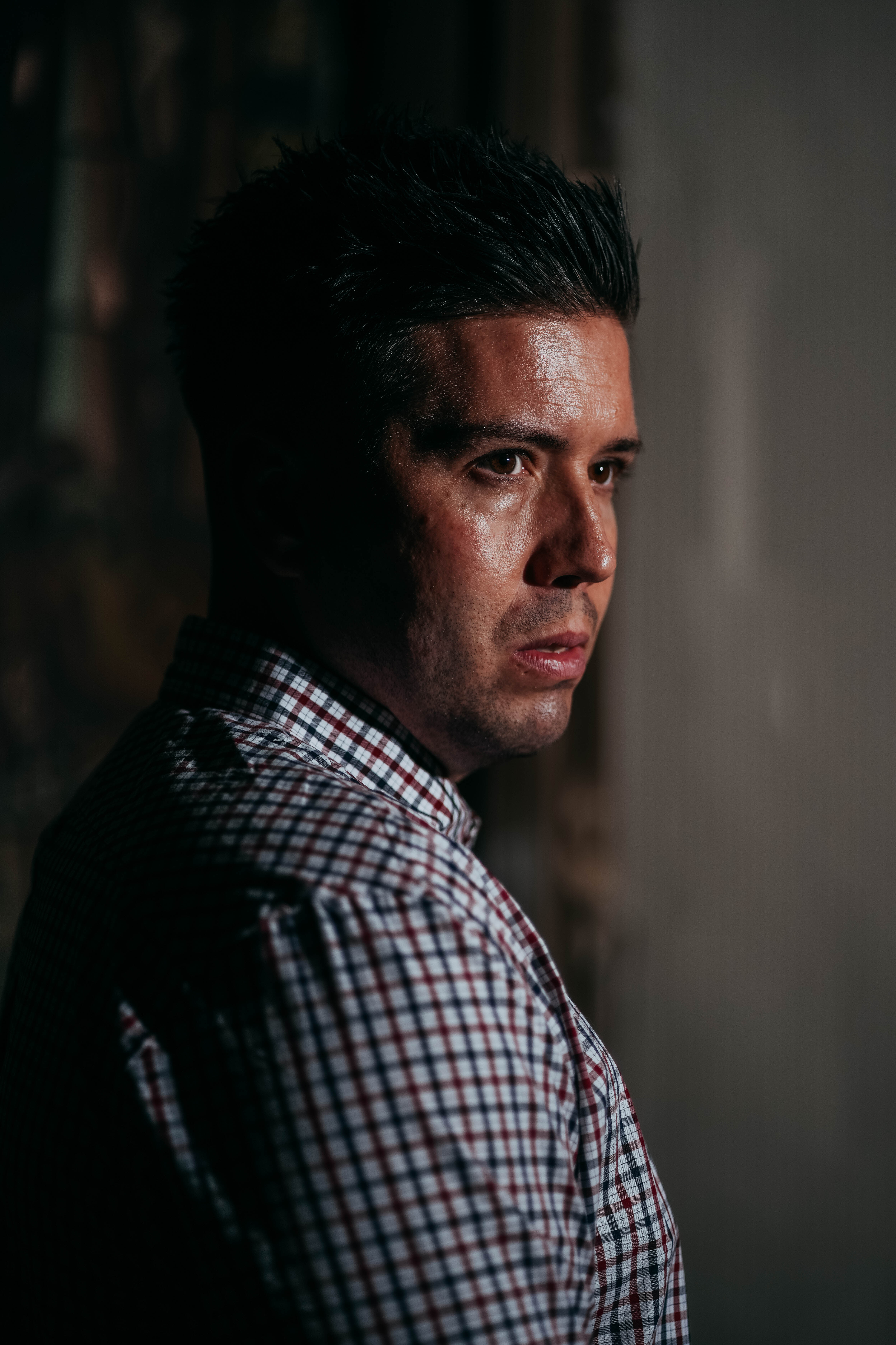 A close-up, dramatic portrait of a man with short, dark hair, wearing a plaid shirt. He is looking over his shoulder, and one side of his face is strongly lit, highlighting his pensive expression against a dark background.