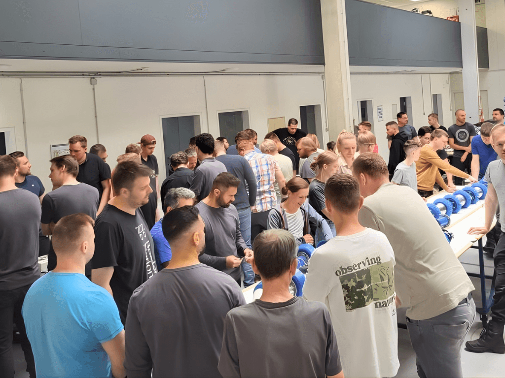 A large group of people gathered, some looking down at a table with blue objects, possibly a demonstration or fair.