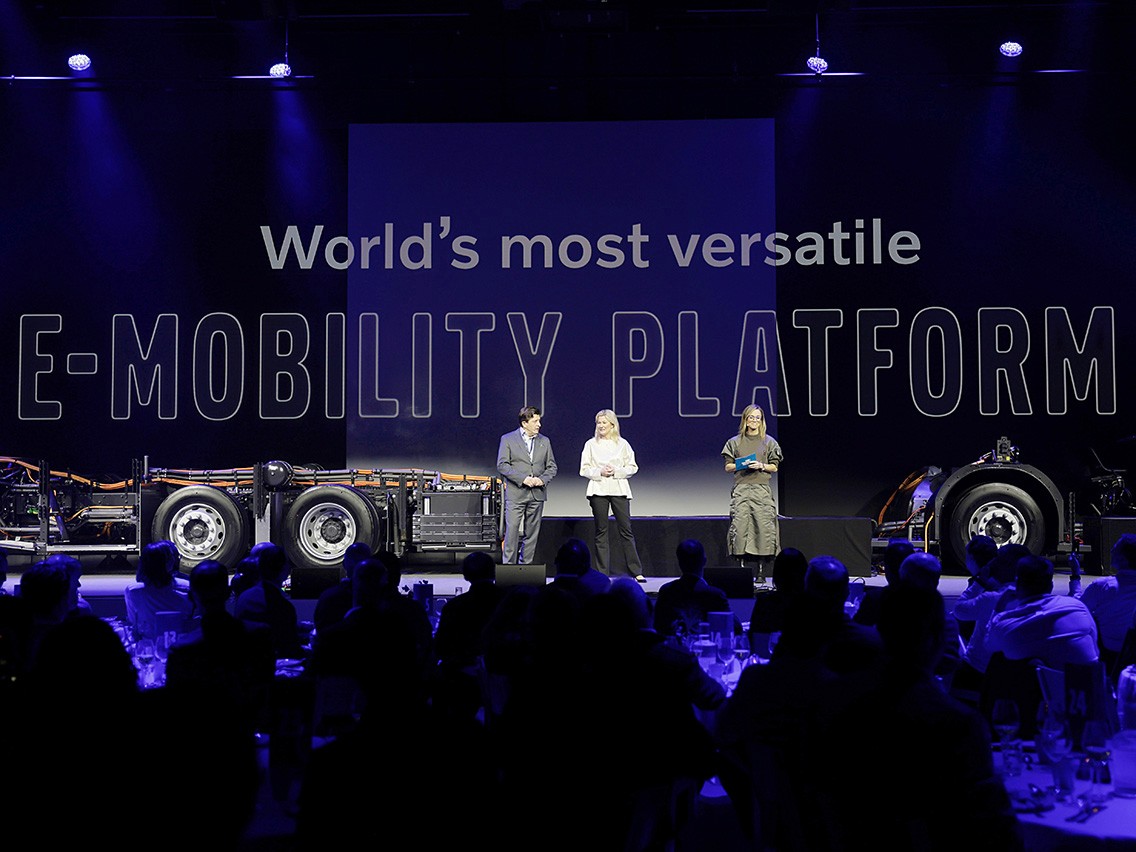 Tre personer står på scenen med en lastbilsplattform framför en stor publik