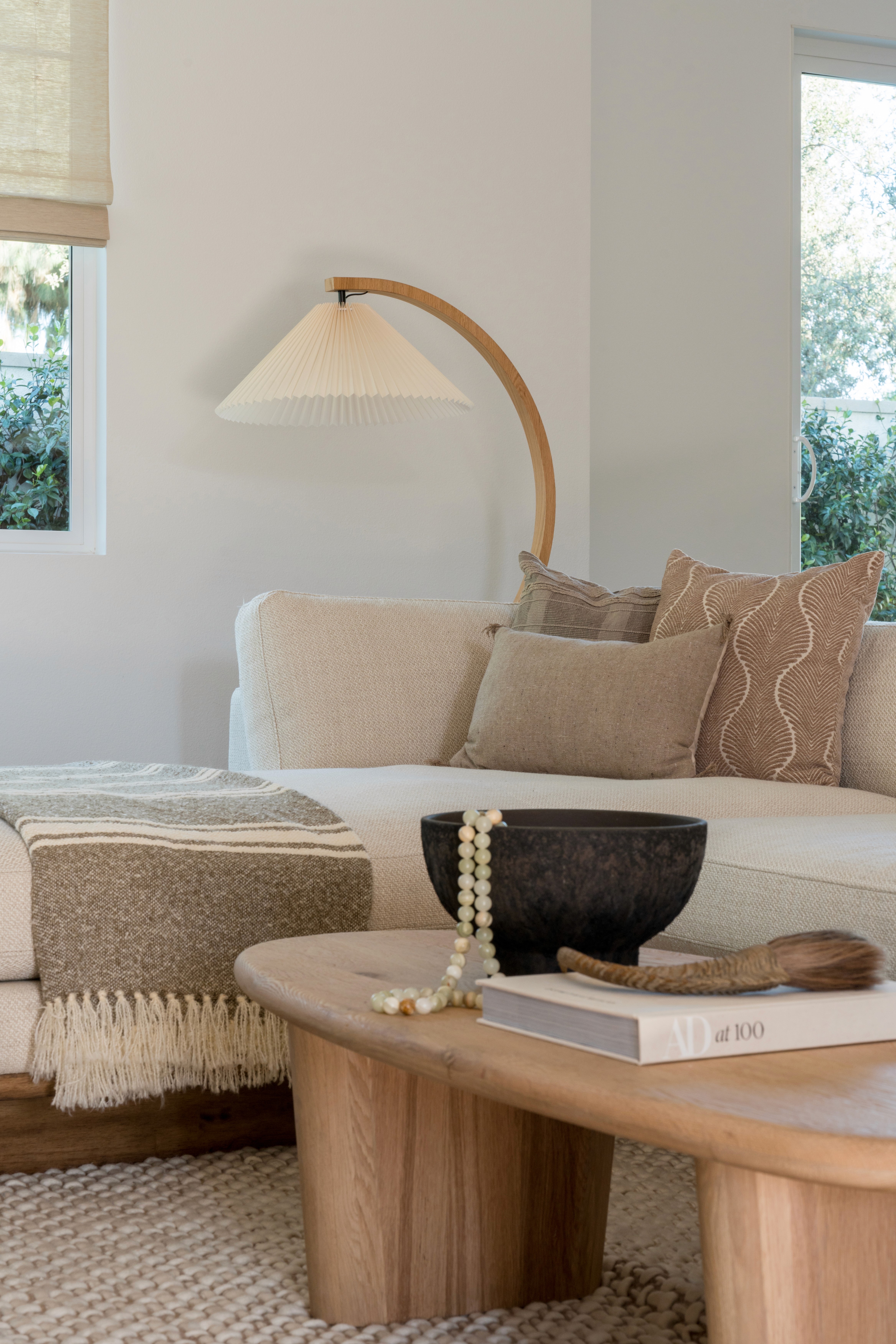 Living room with oval, wood coffee table, pleated floor lamp, and white linen sectional with textured pillows and throw blanket by Jubilee Interiors in Arcadia, California