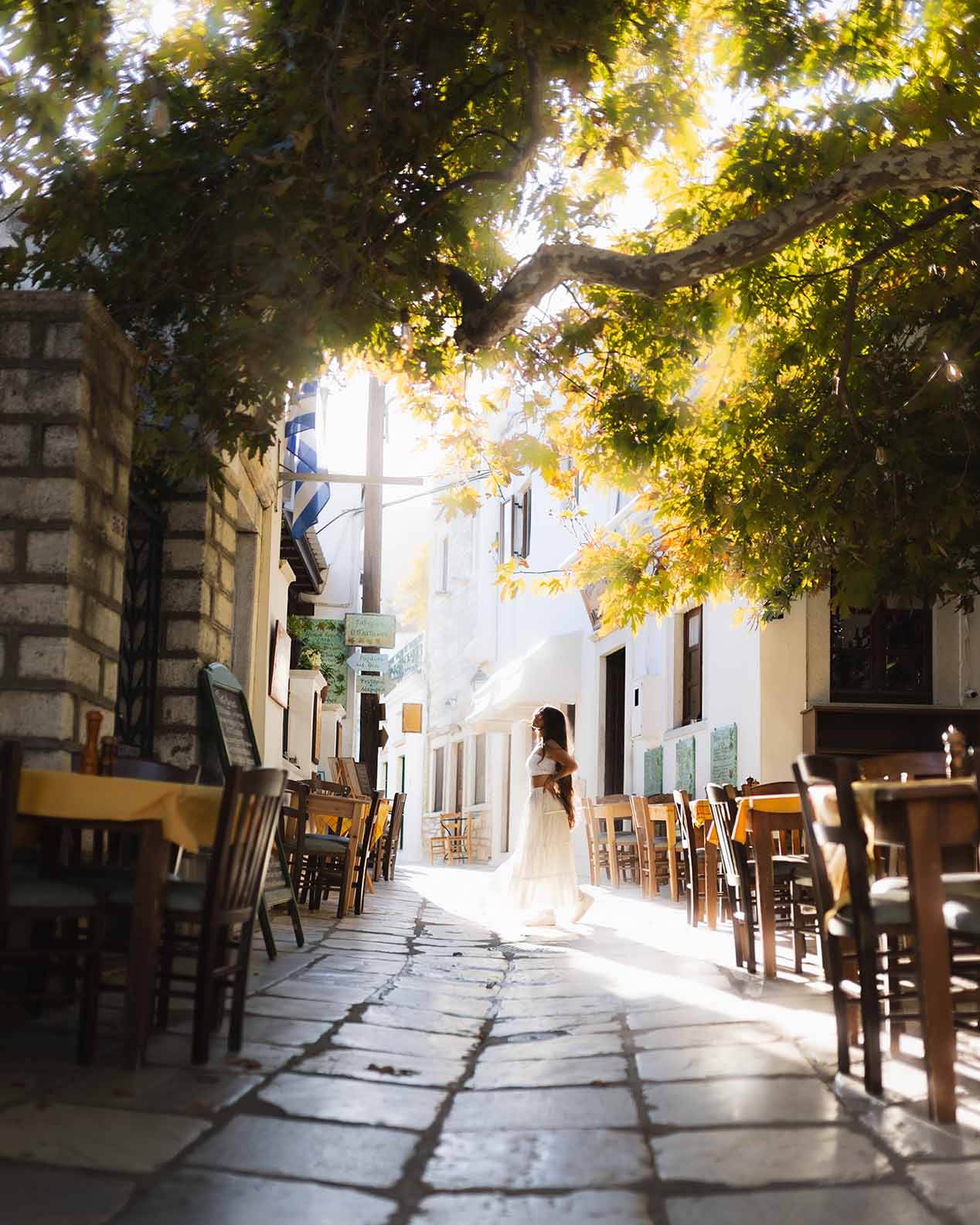 Alleyway with restaurants on each side in Greece, with a girl in a dress standing in the middle in beautiful light.