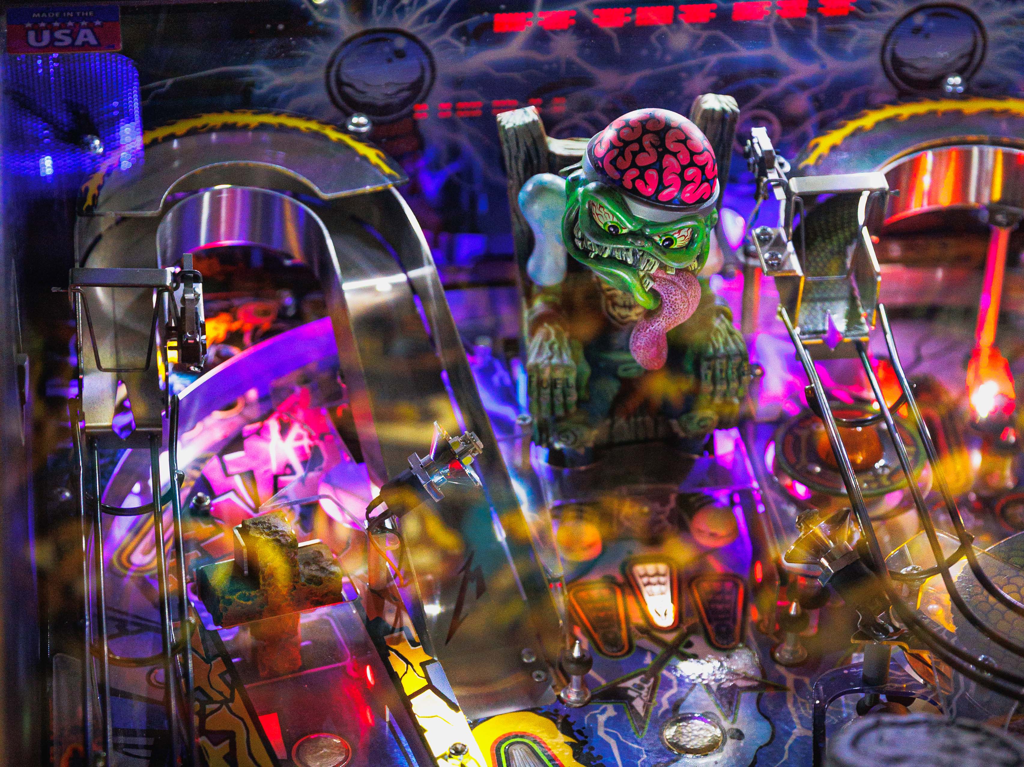 Close-up of the Metallica pinball machine playfield showing a green monster with an exposed brain and long tongue, surrounded by ramps, lights, and colorful artwork.
