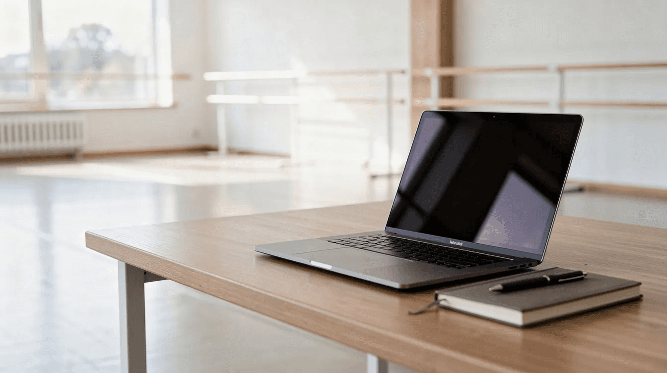 The image shows an open laptop on a neat desk in a bright studio space, with ballet barres in the background, suggesting a professional development environment ready for educators to prepare their classes for September. This setting symbolizes a successful start to the school year, where technology and self-care can align with teaching goals and resources.