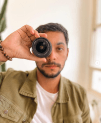Foto de Thiago Almeida, ele segura uma lente de cãmera fotográfica, posicionado-a na frente de seu olho direito.