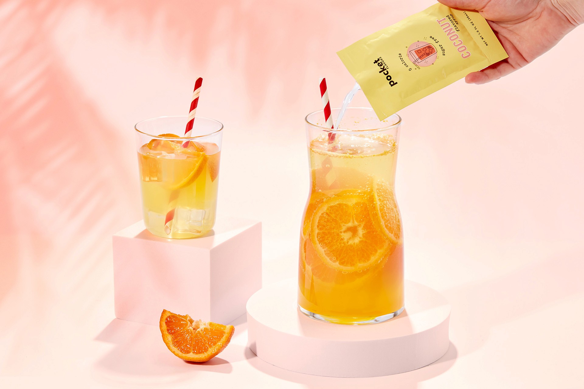 Two glasses of refreshing orange-flavored drinks, each with sliced oranges and red-striped straws, are displayed on white pedestals with a hand pouring Pocket Flavors from a packet on a peach-colored background.