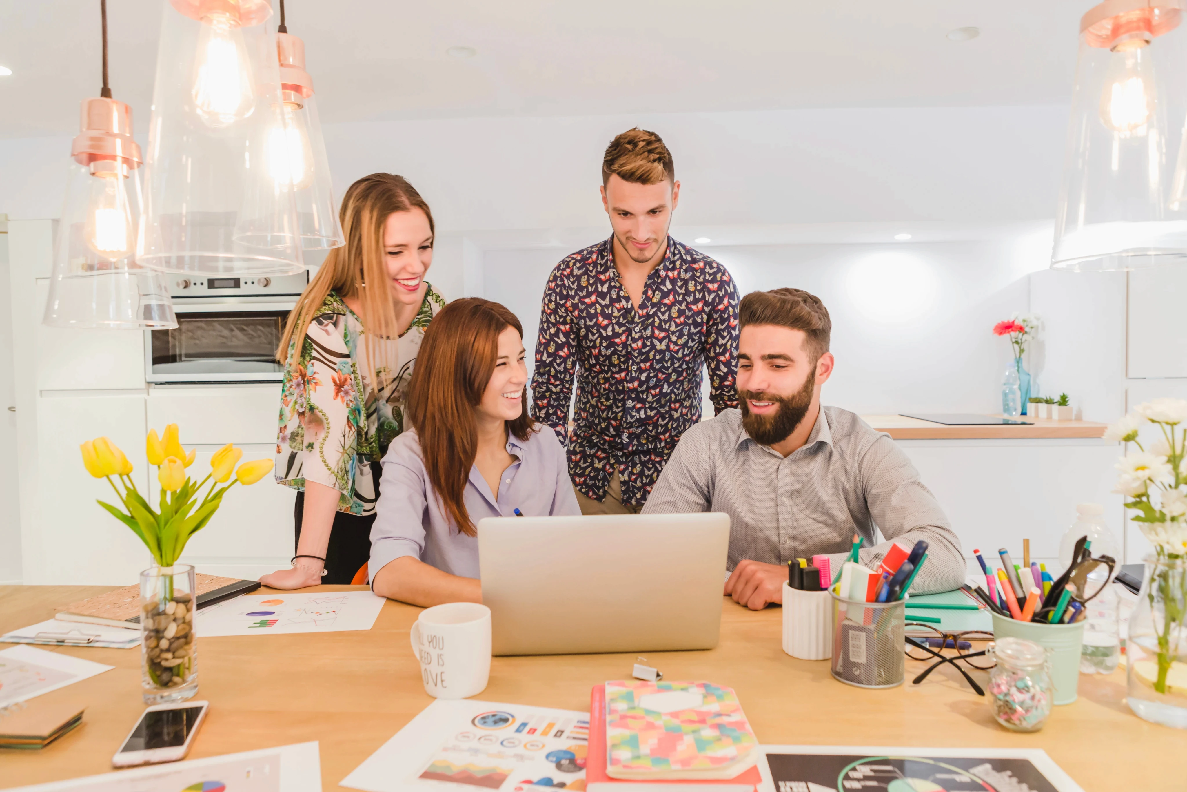 A group of four people smiling and collaborating around a laptop in a bright office. The desk has colorful stationery, and flowers are in the background.