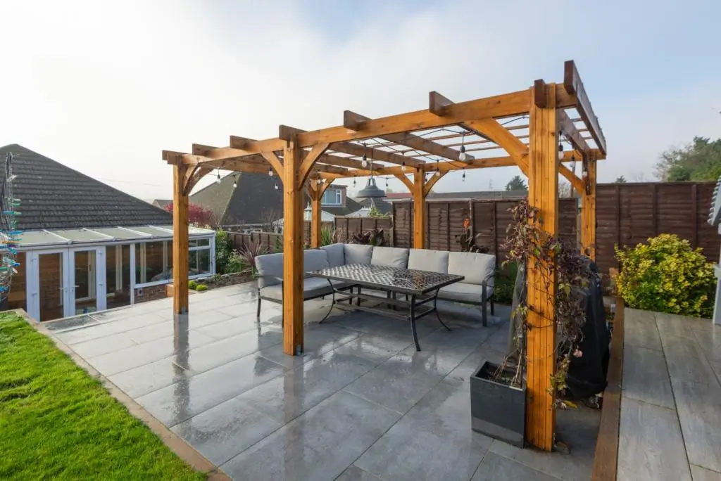 A wooden pergola with a dining set underneath, set on a patio with a lawn and house in the background.