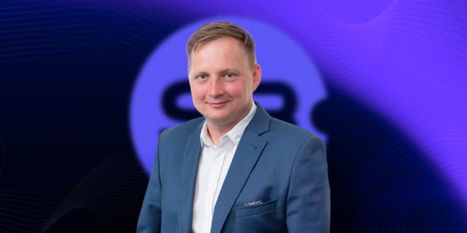 A smiling Michał Głowacki, COO of Sb Software, with short blond hair, wearing a blue blazer and white shirt, looks directly at the camera. A blurred purple logo is in the background.