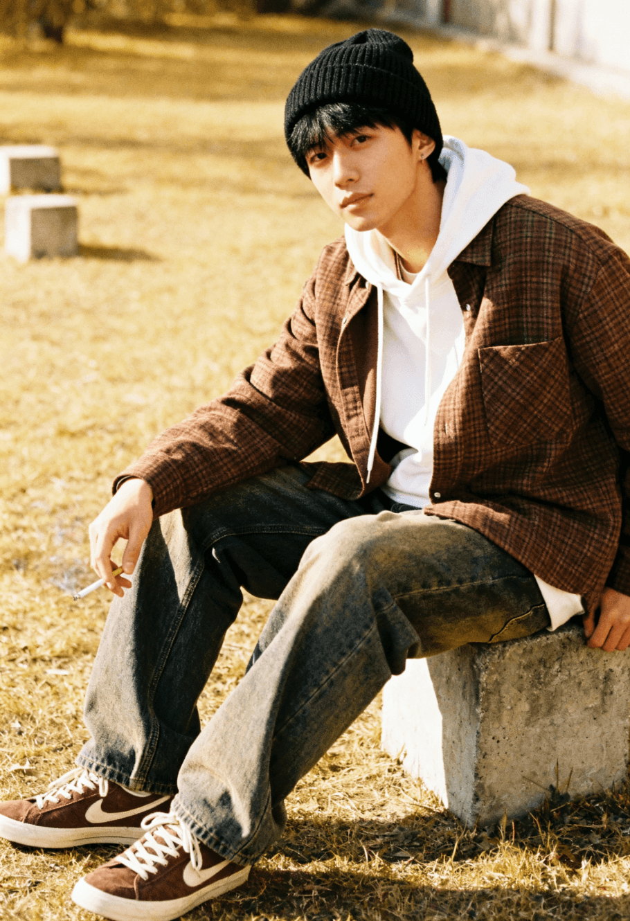 Person wearing a black knit beanie, white hoodie under a brown checkered jacket, and baggy jeans sitting on a concrete block in a sunlit outdoor area with brown and white sneakers