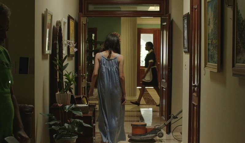 A photo of a woman with a blue dress walking in a hall way while another woman dressed as a maid is walking by.