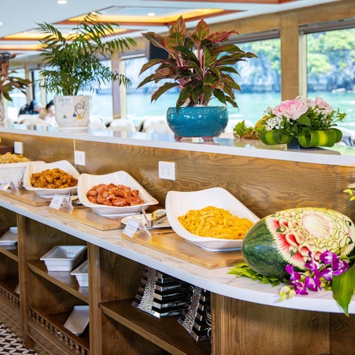 Et buffetbord med forskellige retter, dekorative planter og en udskåret vandmelon, placeret i et godt oplyst spiseområde med naturskønne vinduesudsigter.