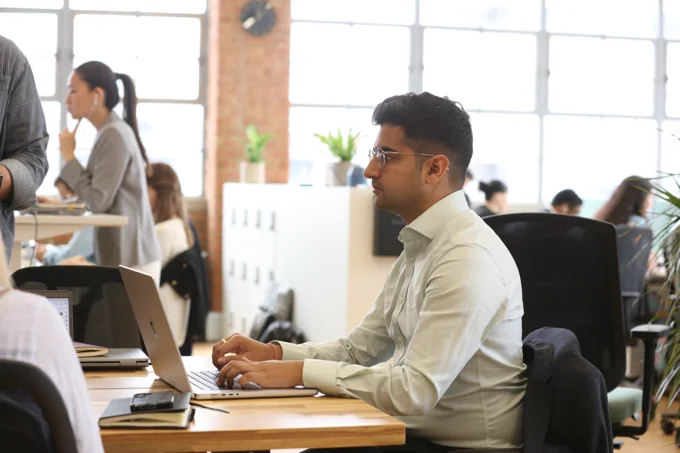 Professional working on a laptop at a desk in an open-plan office with colleagues collaborating in the background.