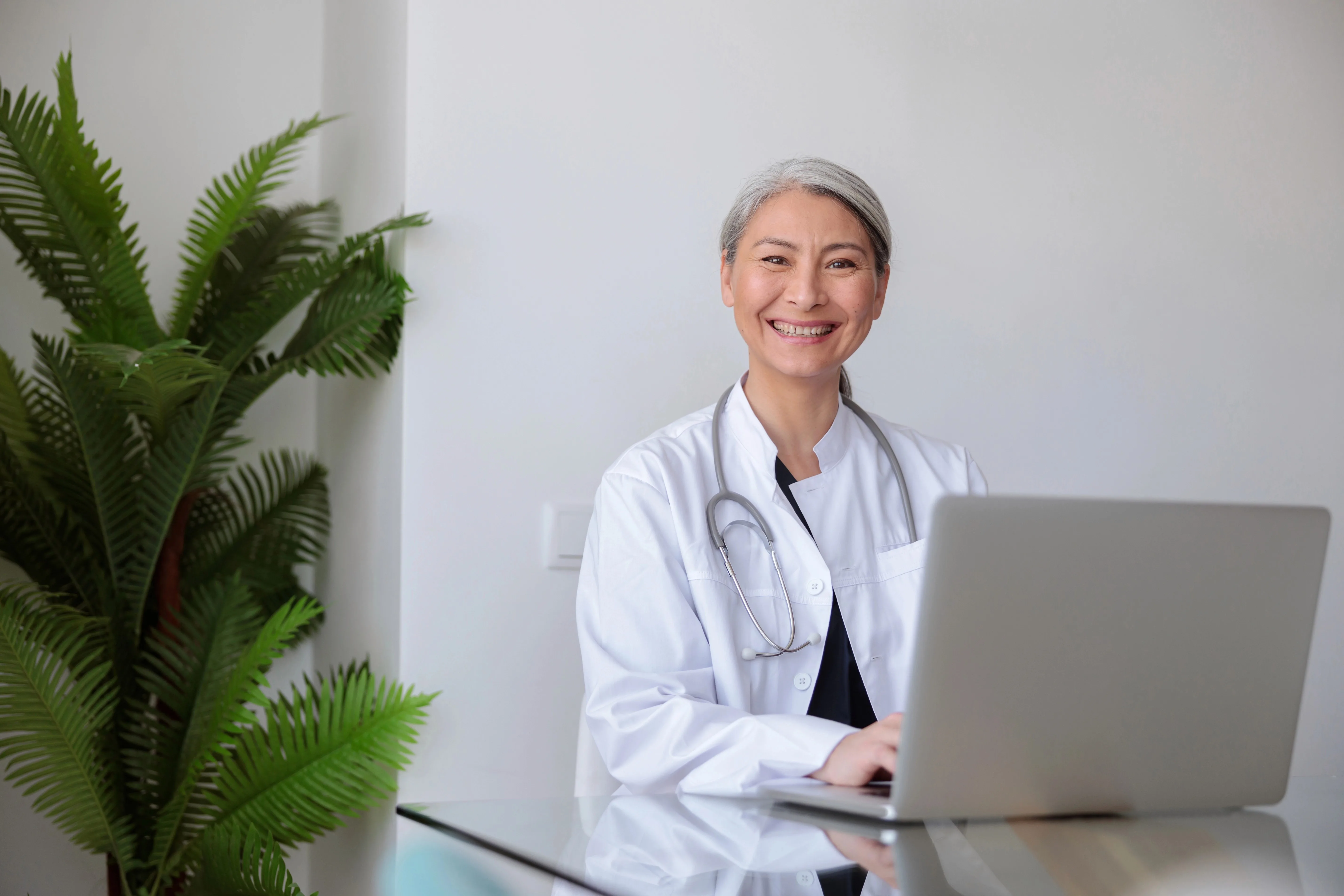 Middle-aged Asian female doctor, sitting at desk with laptop open, smiling at the camera.