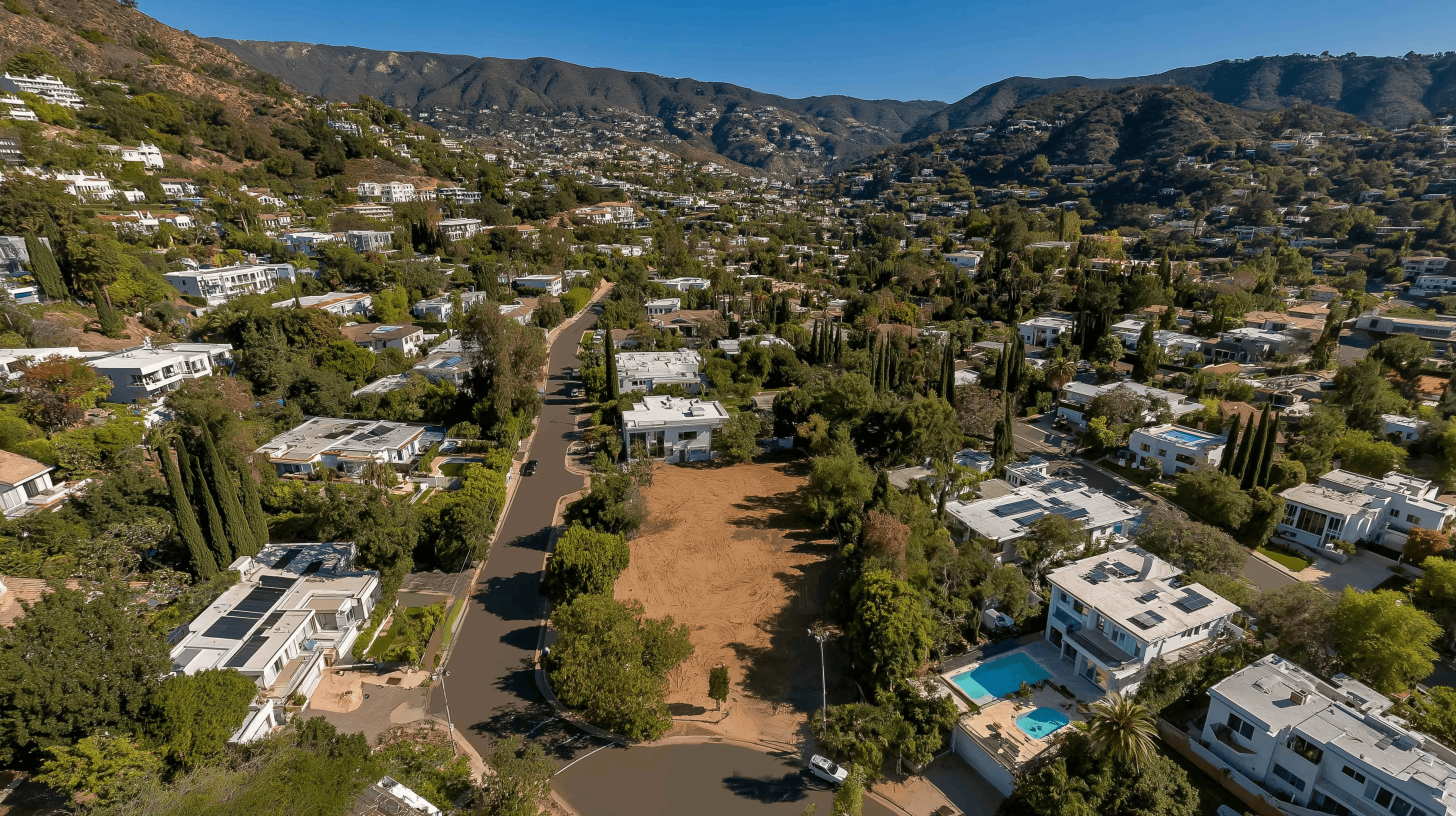 Aerial view of a sprawling hillside neighborhood densely packed with trees and luxury houses, highlighting a large, cleared dirt lot in the center.