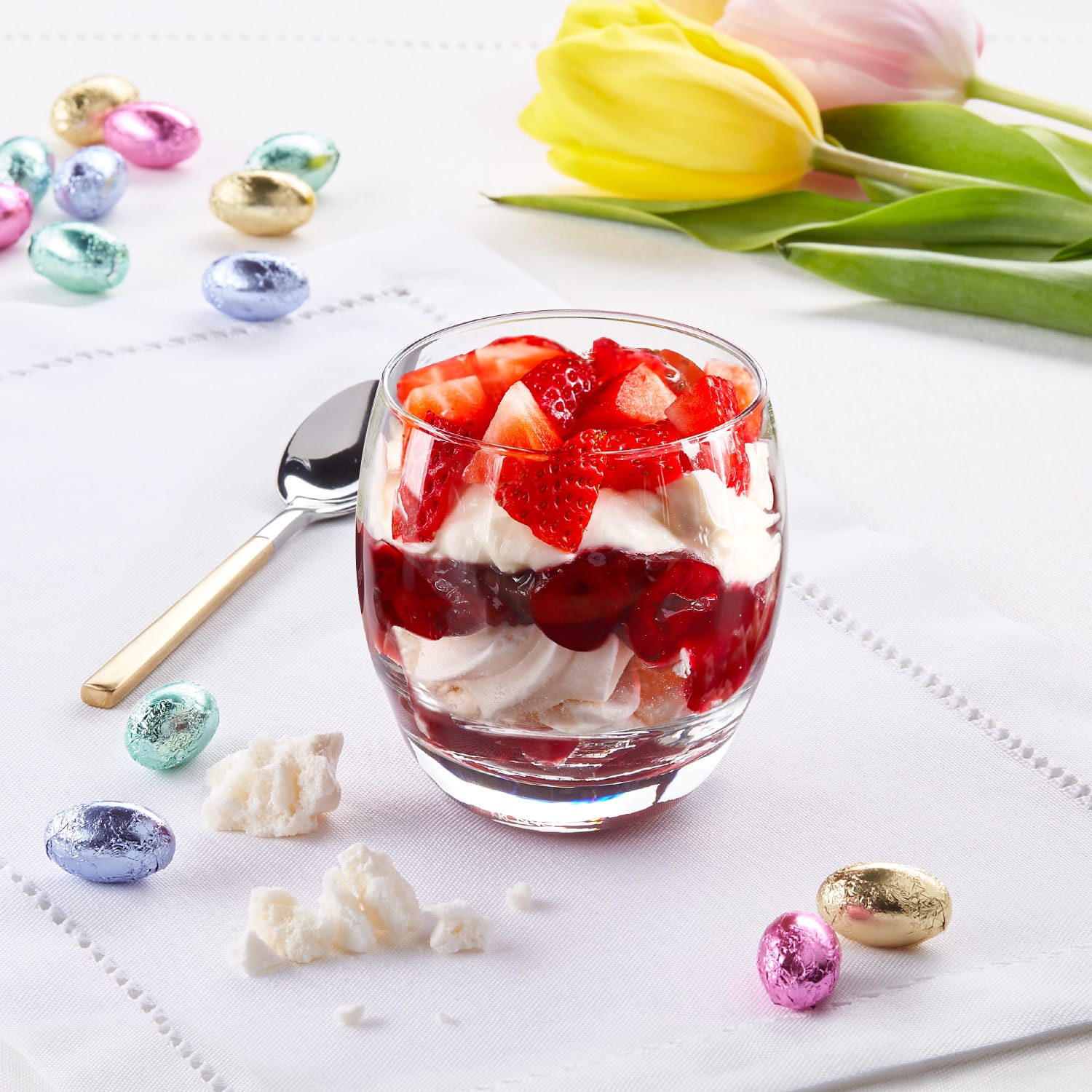 A glass cup filled with layers of fresh strawberries, cream, and jelly sits on a white tablecloth, surrounded by colorful Easter eggs and a yellow tulip in the background.