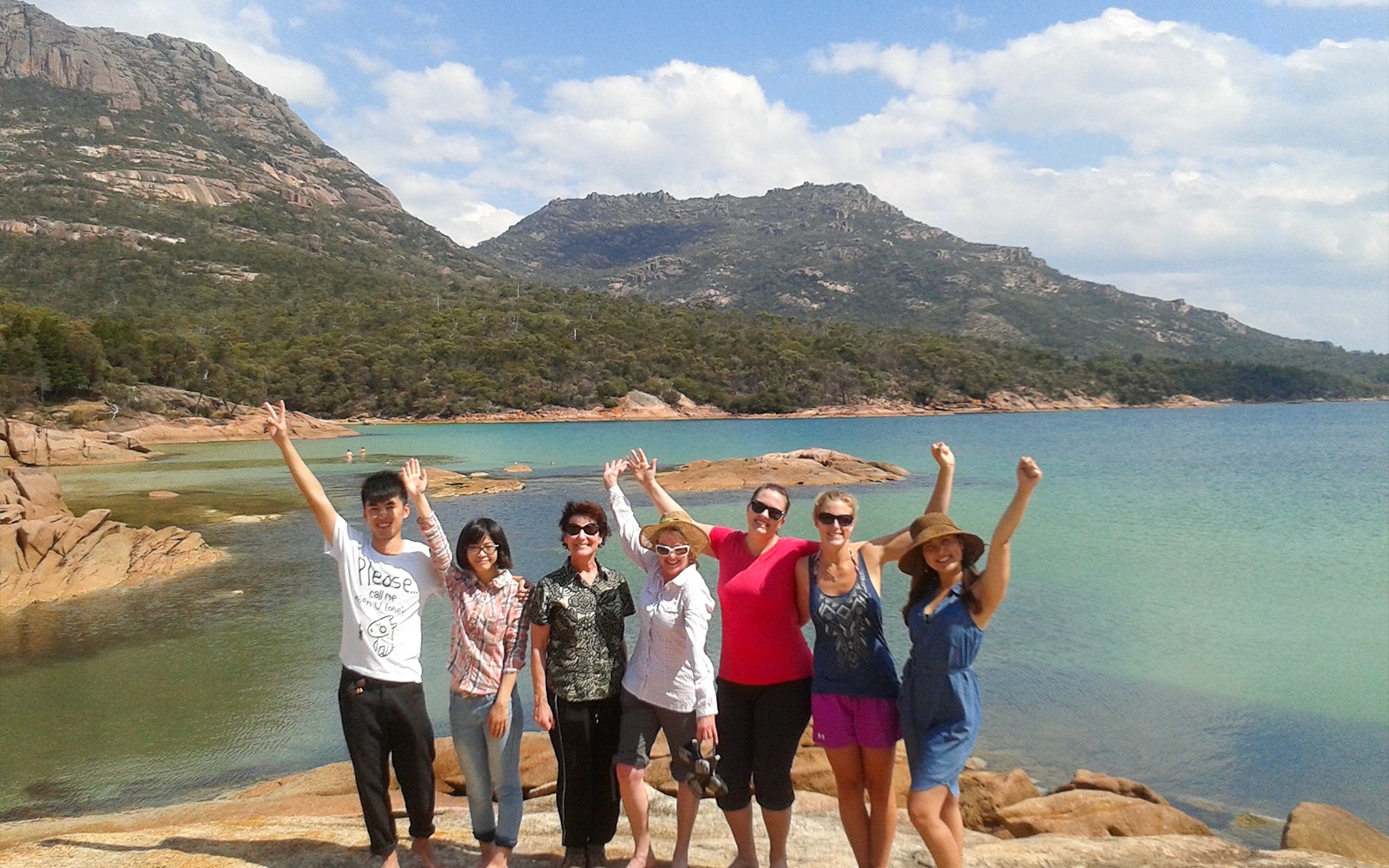 一群遊客在塔斯馬尼亞的酒杯灣欣賞風景，背景中有山脈和清澈的水。
