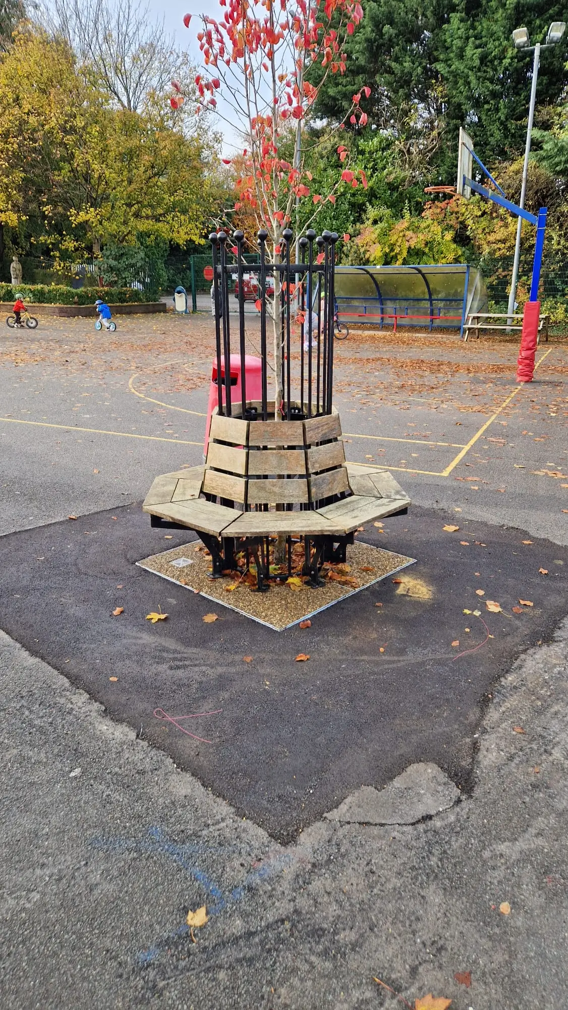 A wooden circular platform with a metal receptacle on top, set on a paved area surrounded by trees.