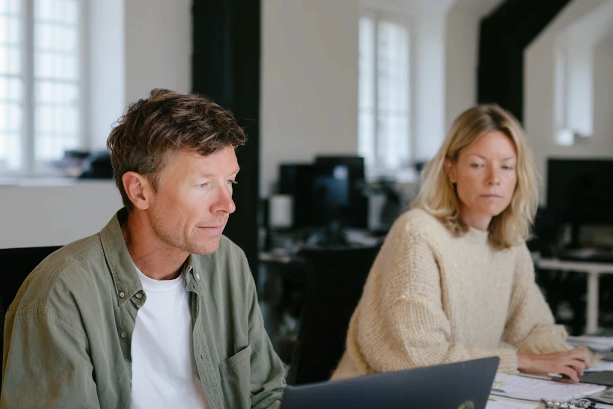 A man and woman engaged in a conversation at a desk with laptops in a modern office setting.