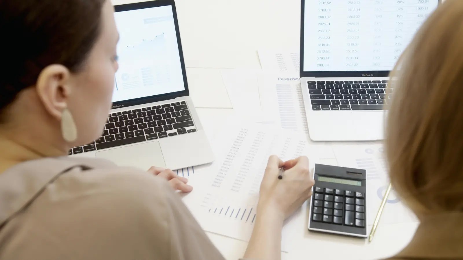 Two people are sitting at a desk analyzing financial documents with spreadsheets displayed on laptops and a calculator positioned nearby, focusing on business data analysis and financial planning with Rockhopper software which simplifies spreadsheets.
