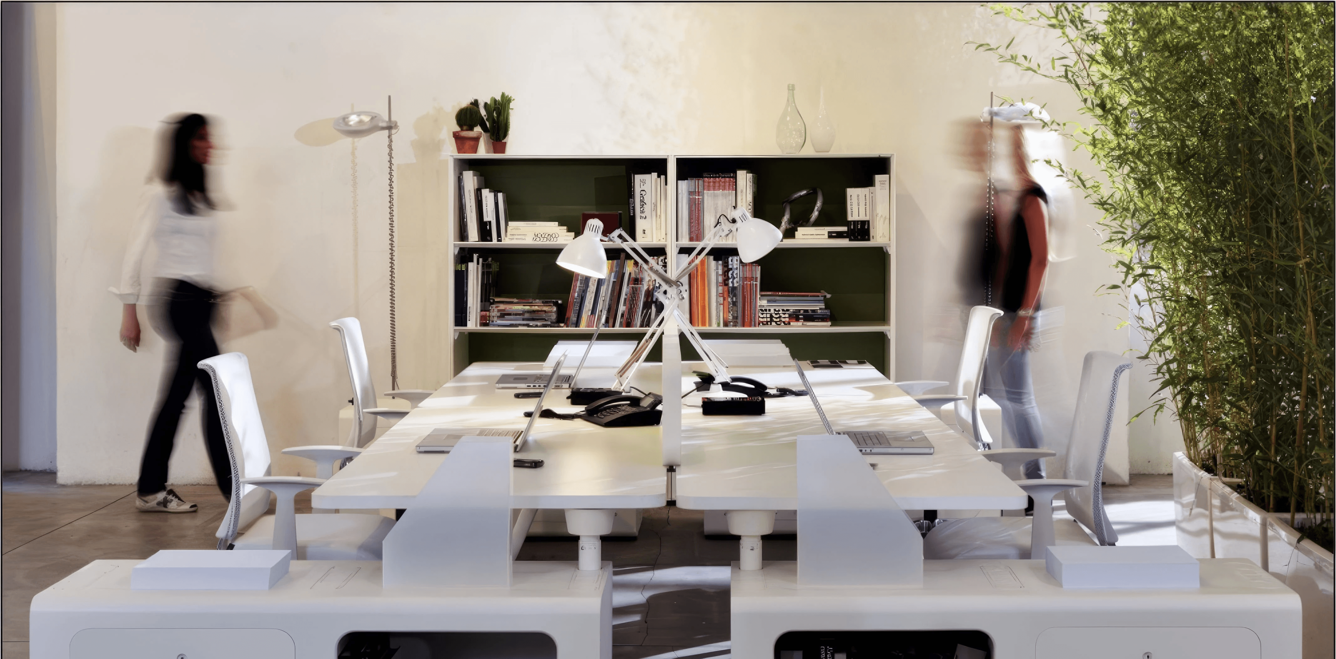 MW Interior - Modernes weißes Büro-Bench-System mit integriertem Unterbauregal, weißen Drehstühlen und Bücherregal im Hintergrund" Beschreibung: Durchdachtes Bench-Arbeitsplatzsystem in Reinweiß mit integriertem offenem Unterbauregal für Bücher und Ablage – funktional kompakt, gestalterisch konsequent und mit zwei Mitarbeiterinnen in bewegter Atmosphäre inszeniert.