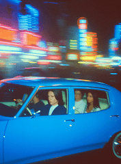 A blue car with people inside is parked in front of colorful neon lights at night.