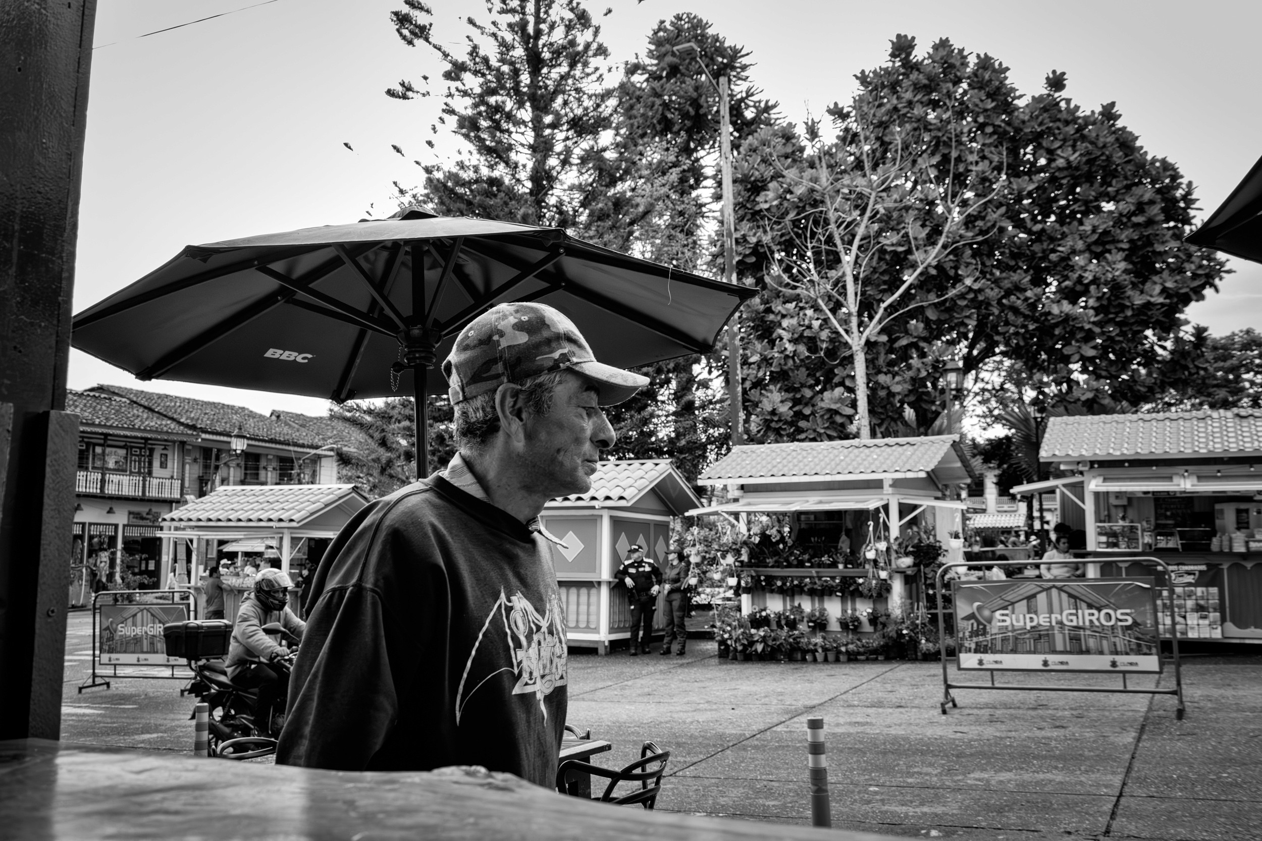 A person wearing a cap stands near a street vendor, with vehicles and trees in the background. Black and white image.