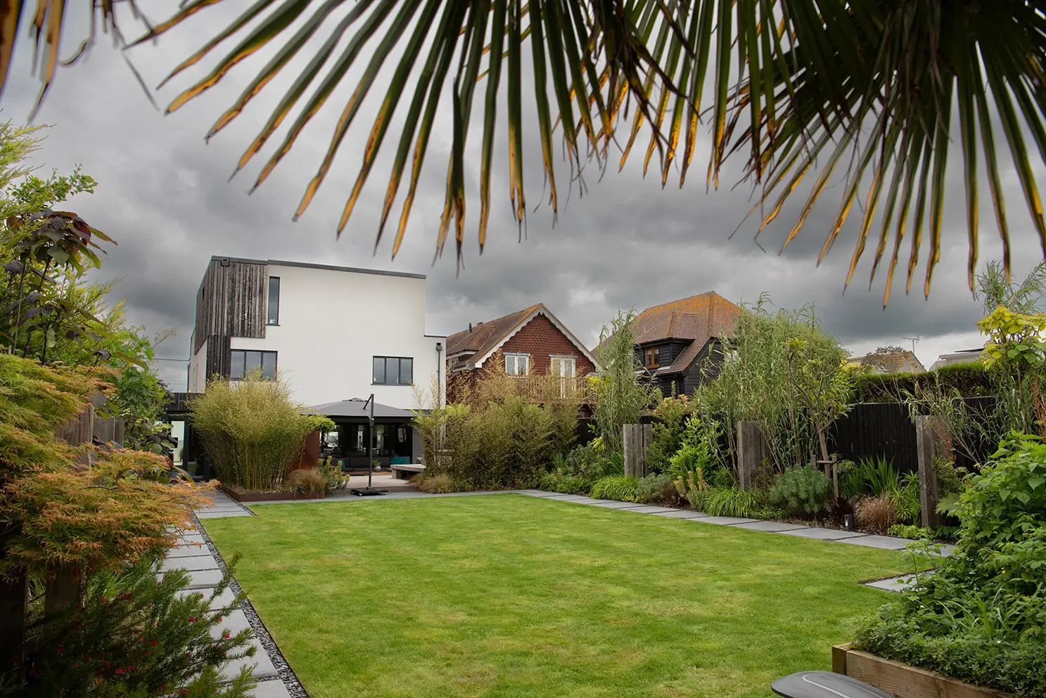 A lush garden with green grass and tropical plants, featuring houses in the background under cloudy skies.