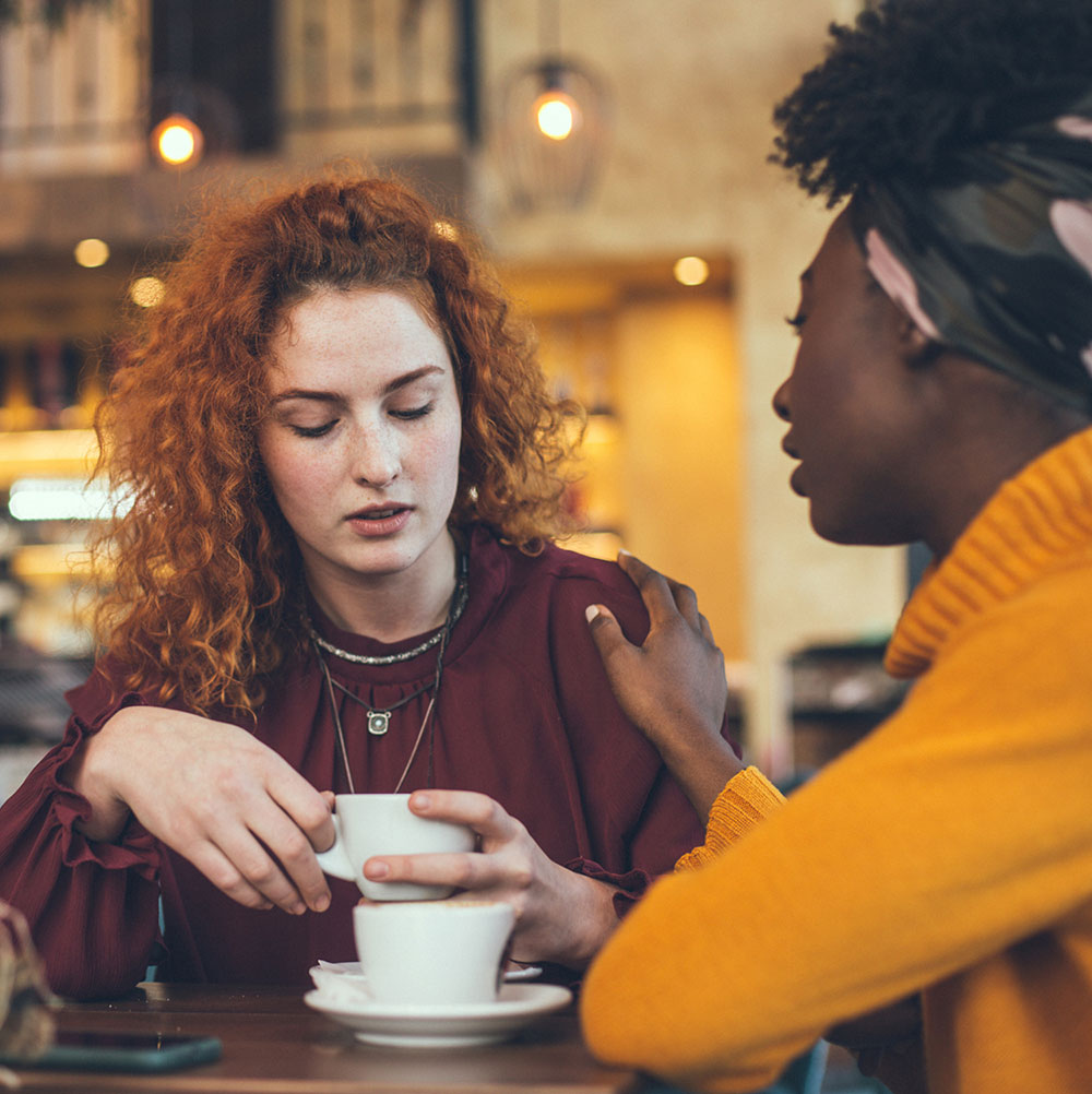 A woman with curly red hair wearing a burgundy top looks down at her coffee cup while a colleague in a yellow sweater places a comforting hand on her shoulder. The intimate café setting with warm lighting suggests a supportive conversation between friends or coworkers, depicting emotional support and workplace empathy.