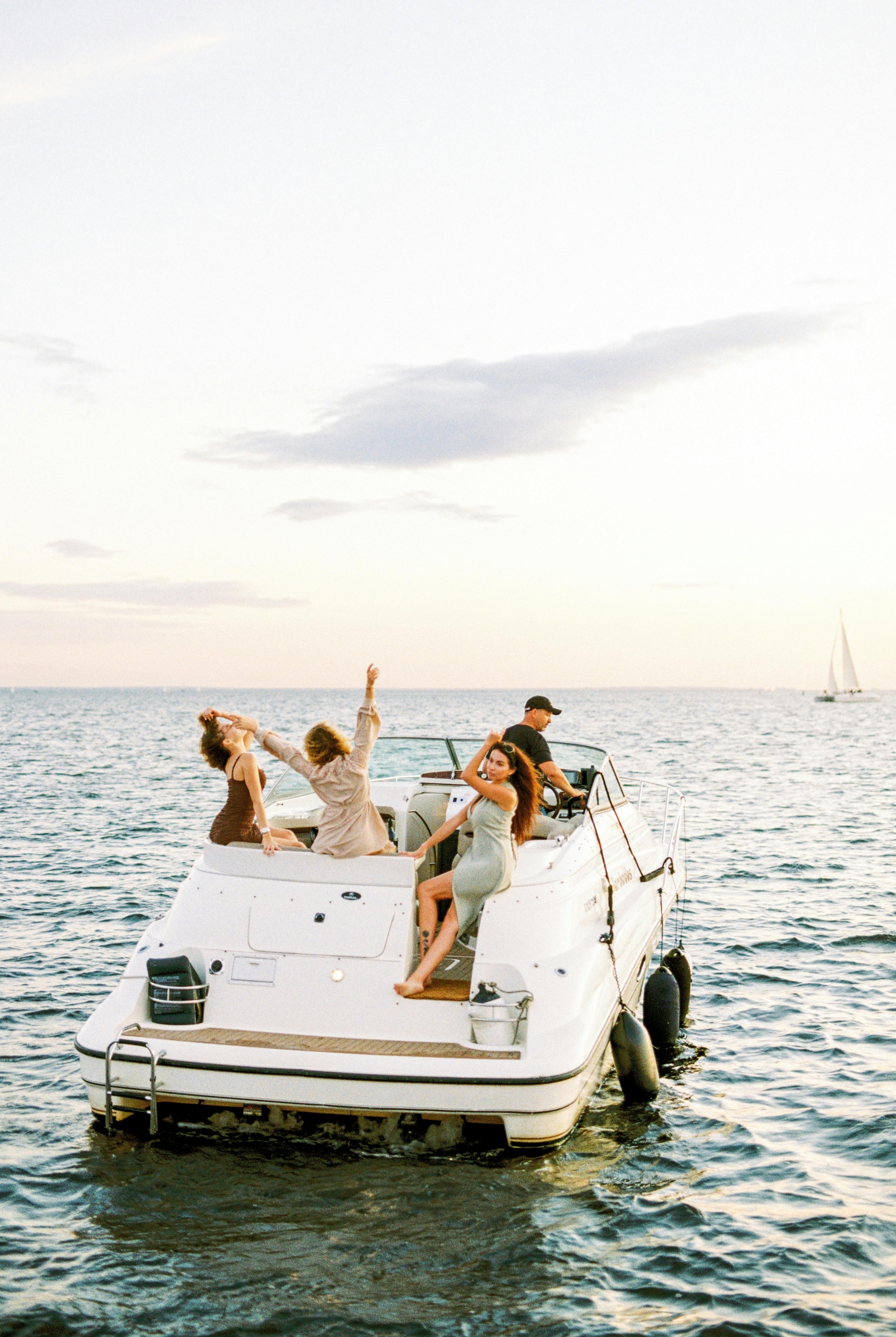 A group of people enjoy a leisurely boat ride on a white motorboat surrounded by calm ocean waters under a clear sky, with one person steering and others sitting and reaching out joyfully, while a distant sailboat is visible on the horizon.
