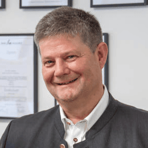 Hubert Zeltsperger steht im Anzug vor einer Wand mit Zertifikaten und lächelt