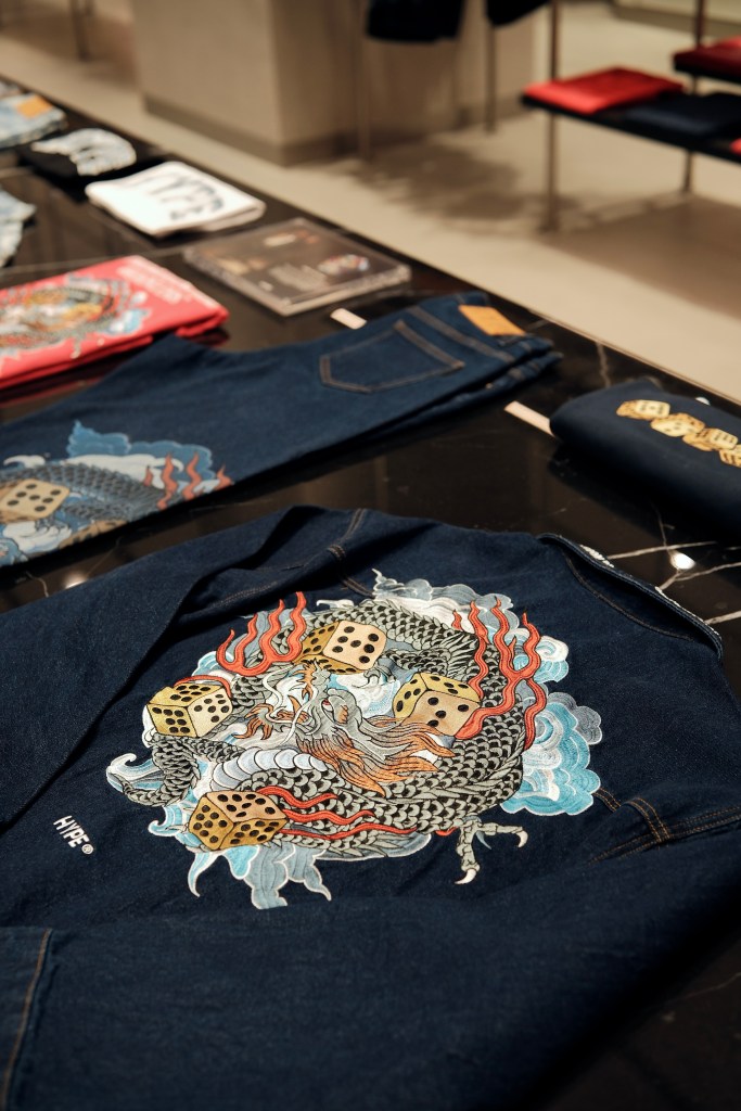Close-up of a denim jacket featuring a dragon design with dice, displayed on a table with other apparel and accessories in the background.