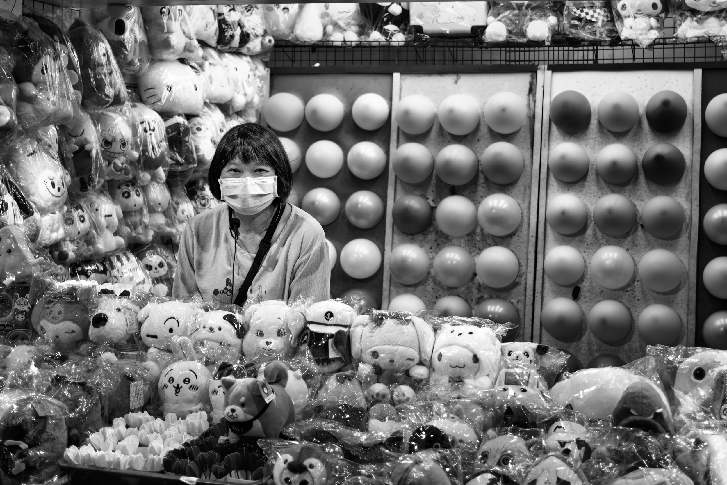 Black and white photograph of a masked vendor surrounded by plush toys at a stuffed animal booth.