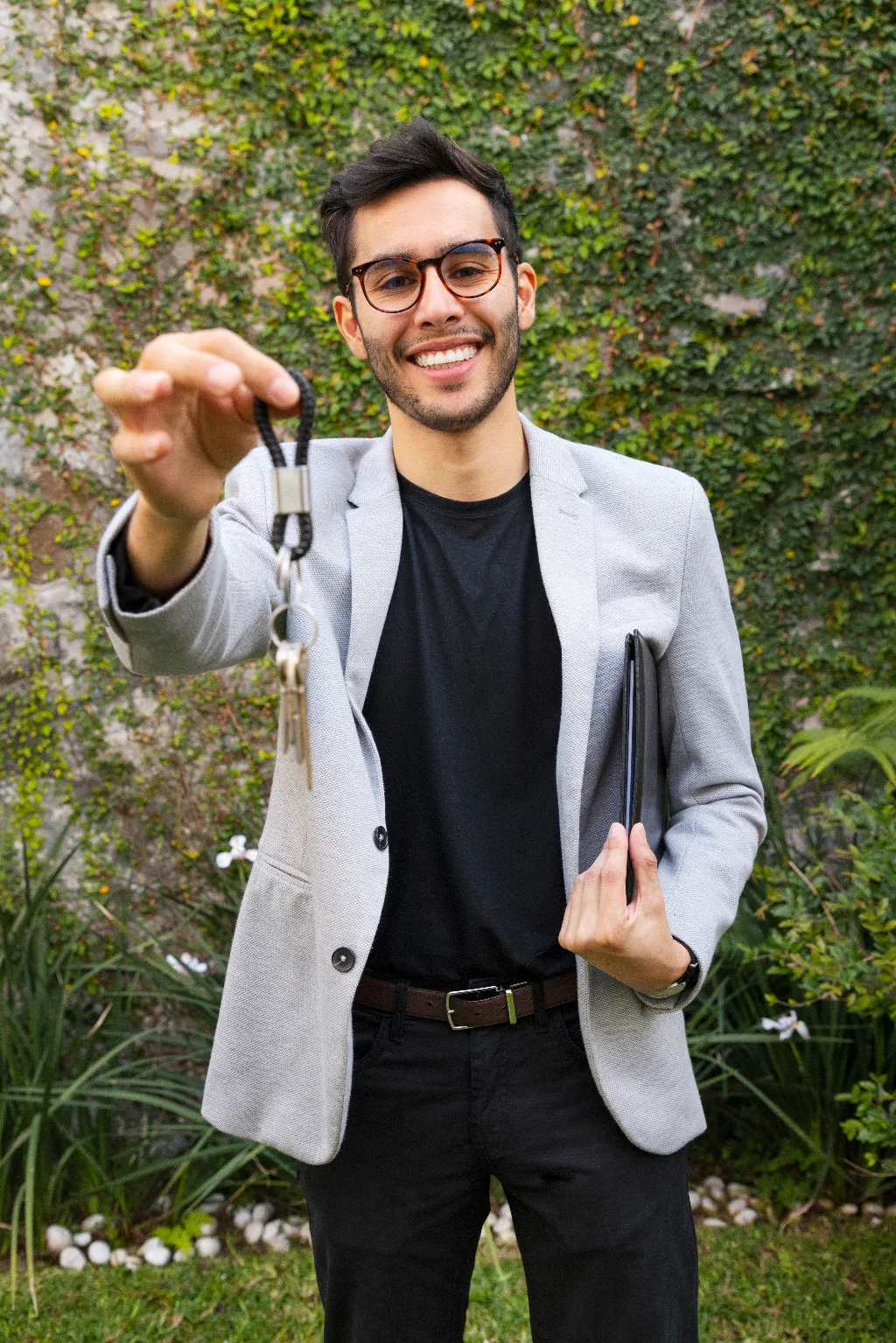 White male standing on a garden, wearing a black shirt with gray coat on top, holding a black folder on the left hand and right hand raised with a key on hand