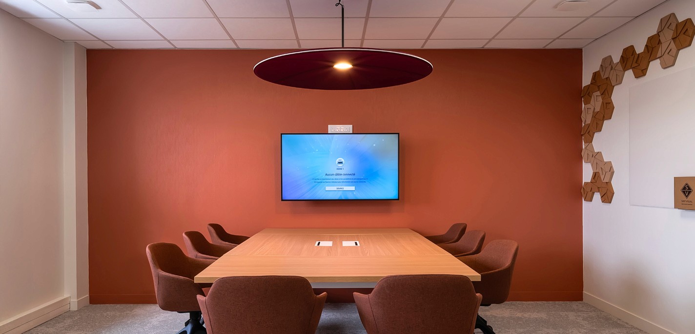 Salle de réunion avec table en bois, écran et chaises marron. Luminaire rond et mur orange – Nord | Photographe pro Anteale | Yann Manac’h