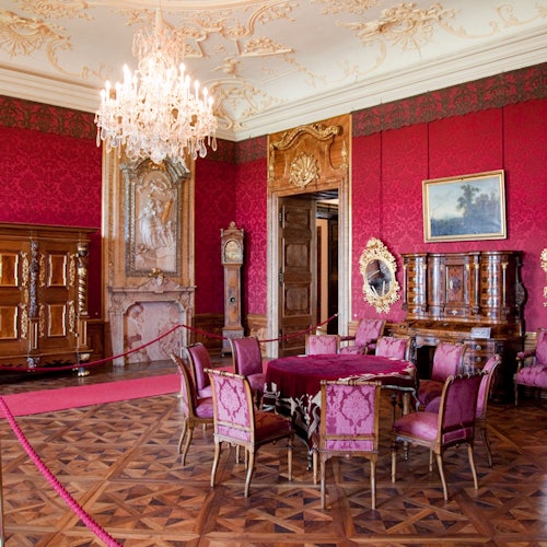 Ornate room with red walls, wooden furnishings, a chandelier, a table with six chairs, a tall clock, and framed artwork.