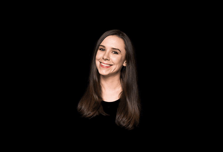 A woman with long brown hair is centered against a solid black background. She is smiling and looking directly at the camera. She is wearing a dark-colored top. The lighting is focused on her face, with no other objects or details visible in the image.