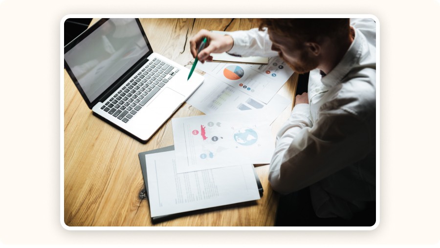 Man analyzes business reports on desk beside laptop and charts.