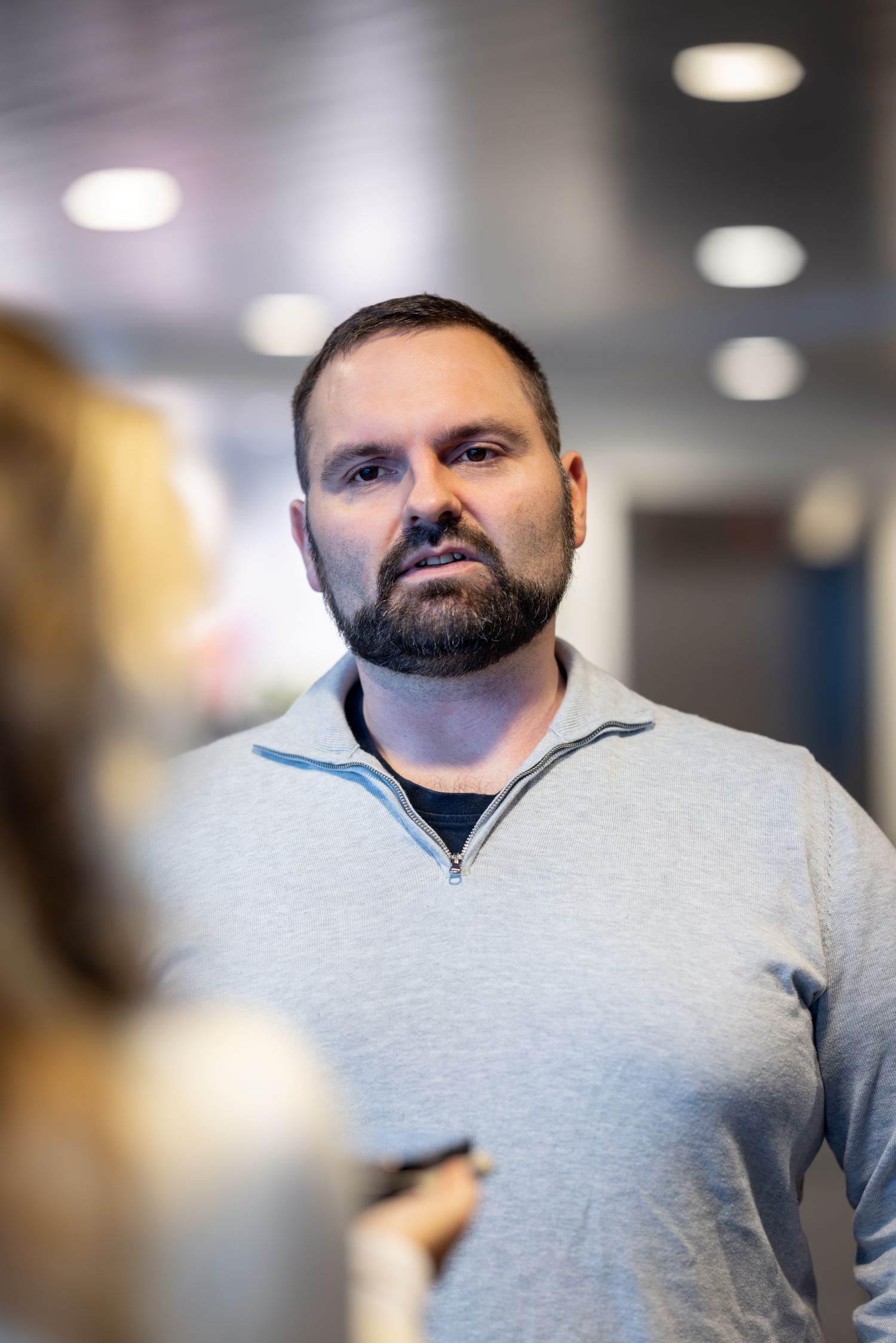 A bearded man in a light sweater stands in an indoor setting, looking slightly away from the camera.