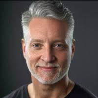 Joel, a white man with grey hair and bear wearing a black shirt and smiling at camera.